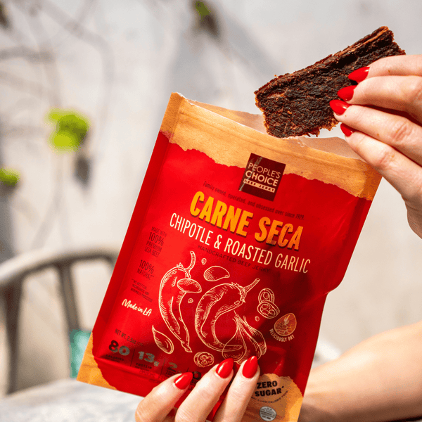 Woman pulling a piece of beef jerky out of an opened bag of Chipotle and Roasted Garlic at a table