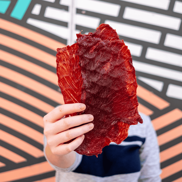 Person holding slabs of beef jerky with a vibrant colored wall behind them. 