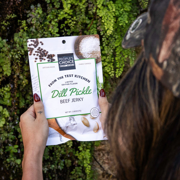 Person holding a package of dill pickle beef jerky in front of green foliage