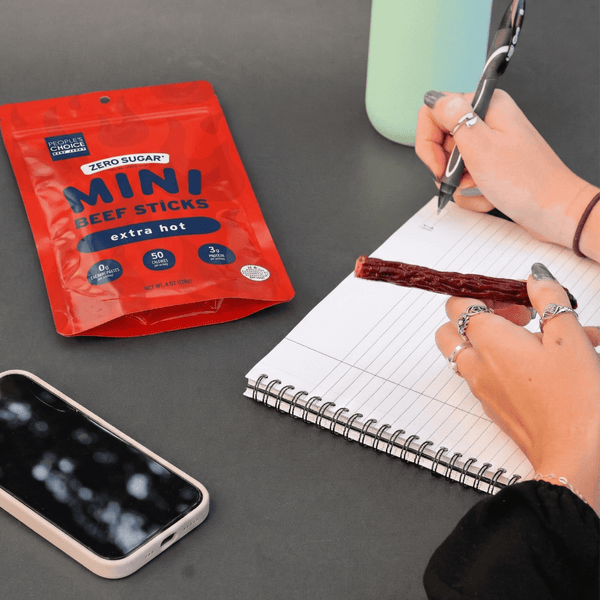 Person eating mini beef sticks next to a packet of 'Zero Sugar' mini beef sticks on a table.