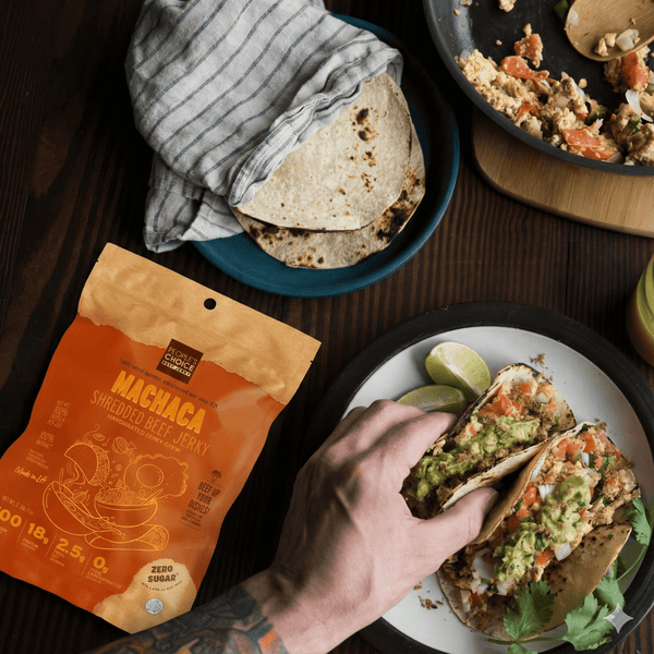 Person holding a taco with a package of Machaca Shredded Beef above a plate of tacos on a wooden table.