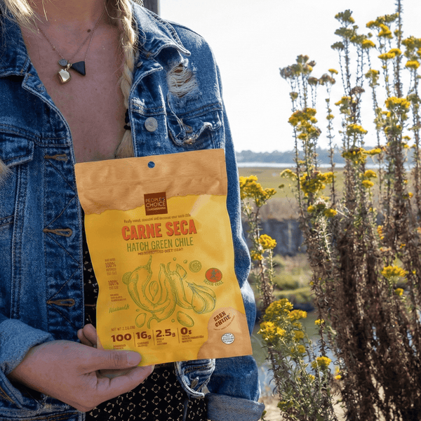 Person holding a bag 'Carne Seca' beef jerky package outdoors with plants in the background