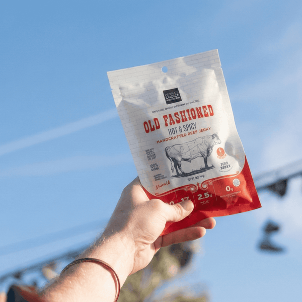 Hand holding a package of Old Fashioned Hot & Spicy Jerky against a blue sky.
