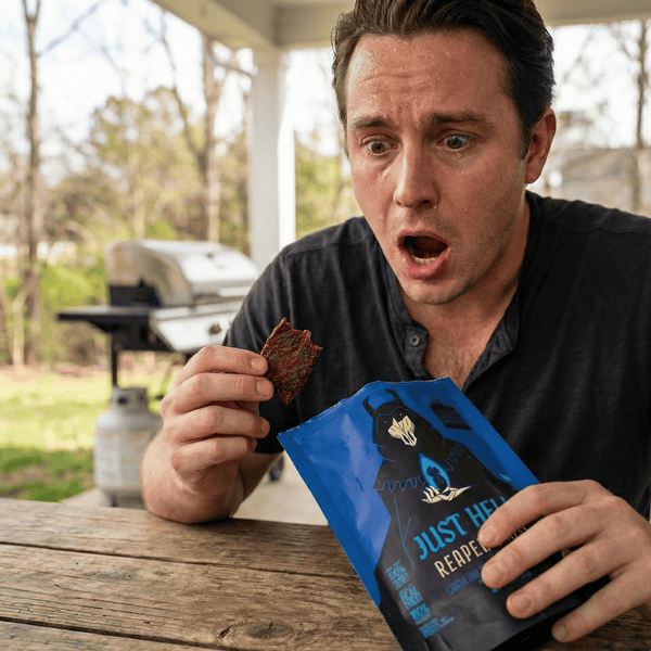 Man holding and eating a package of Just Hell Reaper spicy beef jerky on a patio outdoors