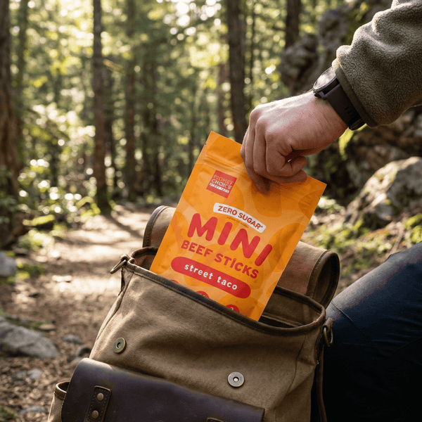 Person placing a package of 'Mini Beef Sticks' into a backpack in a forest setting