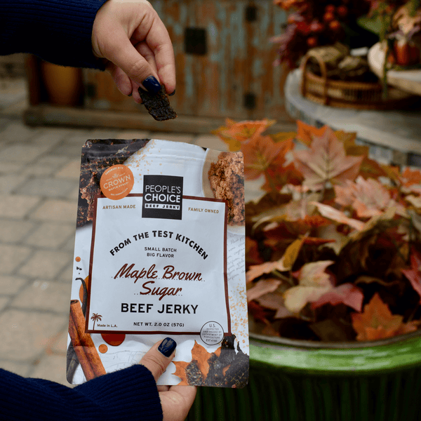 Hand holding a package of People's Choice Maple Brown Sugar Beef Jerky with a blurred autumnal background.