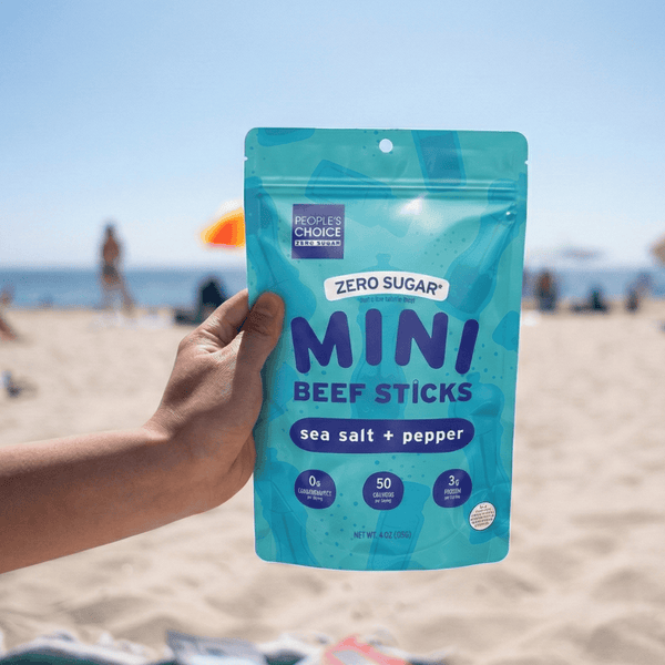 Hand holding a package of Mini Beef Sticks on a beach with people and umbrellas in the background.