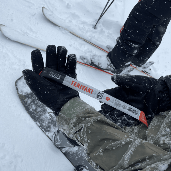 Person holding a 'People's Choice Teriyaki Beef Stick' in snowy conditions with skis in the background.