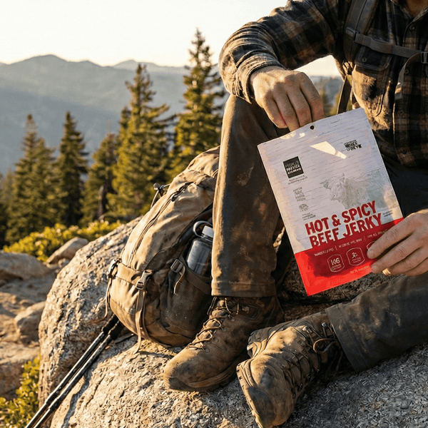 Person sitting on a rock in a mountainous area holding a package of hot and spicy beef jerky.